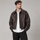 A man wearing a dark brown leather jacket over a white button-down shirt and black trousers, standing against a neutral gray studio background.