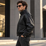 A man wearing a black leather jacket and dark sunglasses walks outdoors in front of a modern building with gray columns.