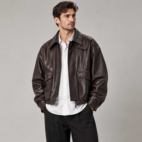 A man wearing a dark brown leather jacket over a white button-down shirt and black trousers, standing against a neutral gray studio background.