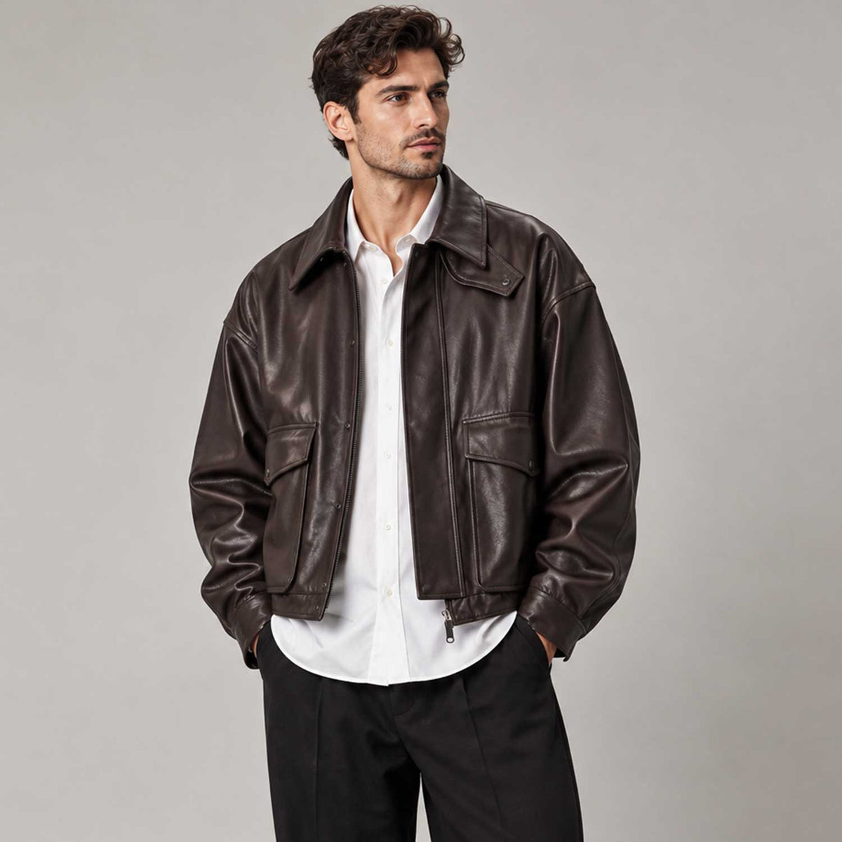 A man wearing a dark brown leather jacket over a white button-down shirt and black trousers, standing against a neutral gray studio background.