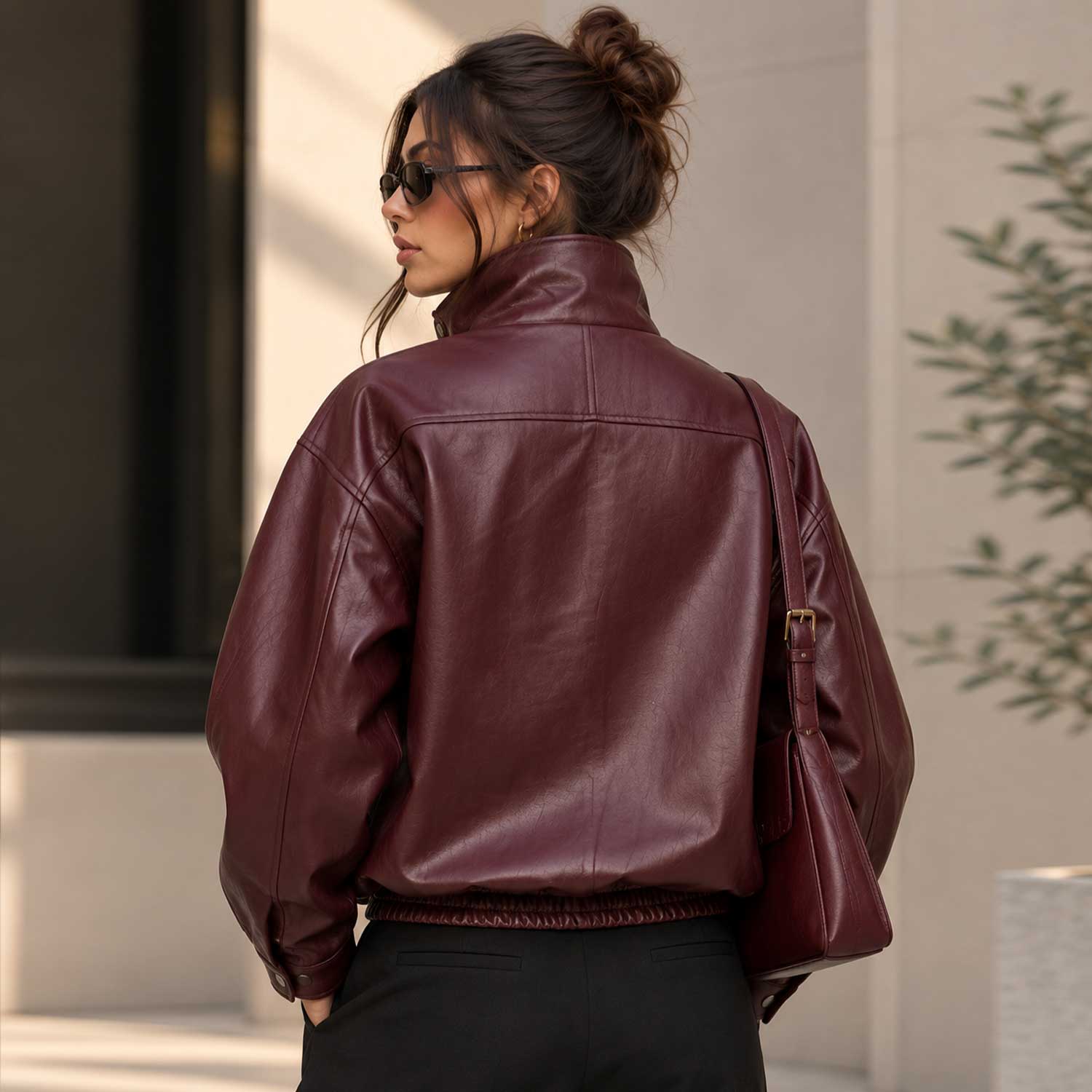 A woman wearing a cropped burgundy faux leather jacket, shown from the back. She is wearing black pants and carrying a matching burgundy shoulder bag in an outdoor urban setting.