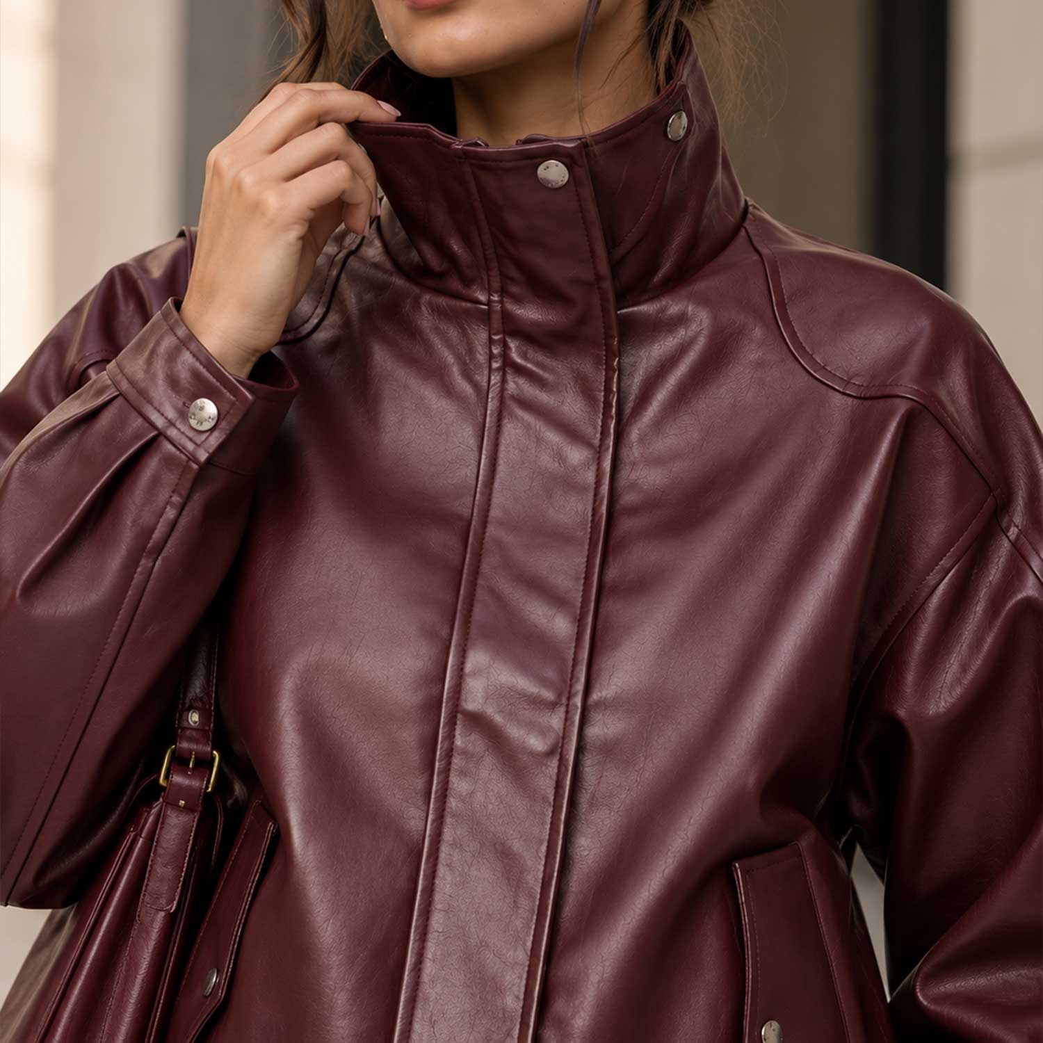 A close-up of a person wearing an oversized burgundy faux-leather jacket with a high stand collar and silver snap buttons. The person is holding the collar up, and a matching burgundy shoulder bag is partially visible.